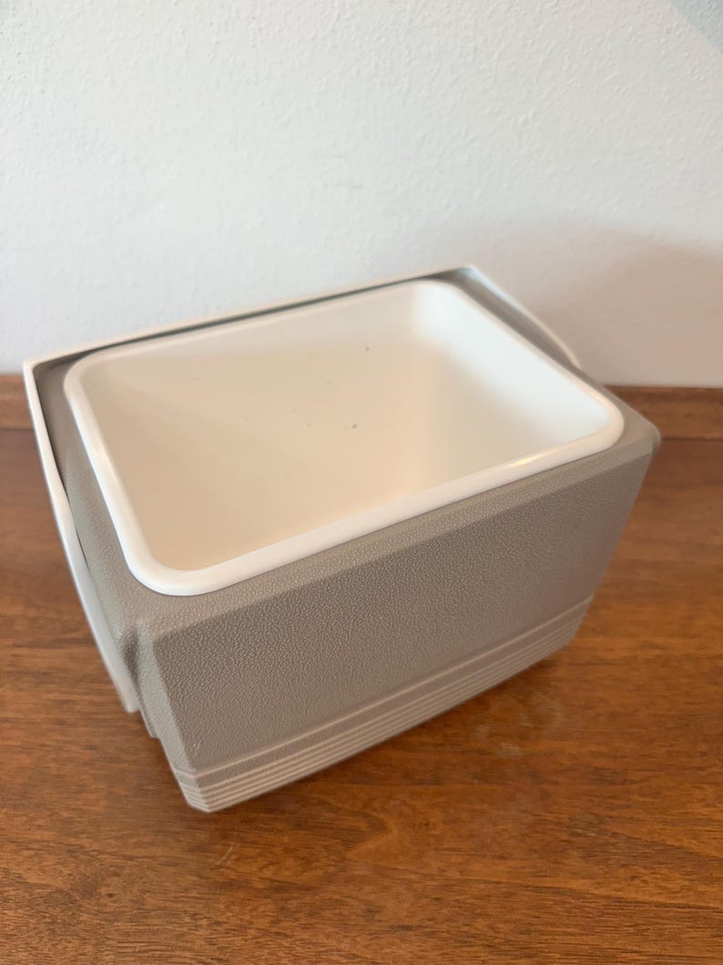 May include: A rectangular ice bucket with a white interior and a grey exterior. The bucket has a textured surface and a white rim. The design is simple and functional, suitable for keeping beverages cold.