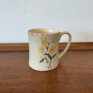 May include: A cream-colored ceramic mug featuring a floral design with yellow daisies and a bee. The mug has a rounded shape with a handle and a slightly textured rim. The design is printed on the front of the mug.