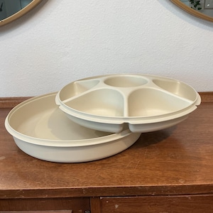 May include: A round, cream-coloured plastic serving tray with a removable insert. The insert features a central circular compartment surrounded by five smaller, wedge-shaped sections, while the base tray is a simple, shallow, circular container.