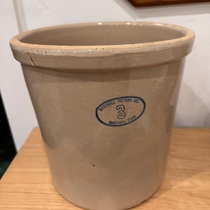 Puede incluir: Una vasija de gres beige de forma cilíndrica con un borde grueso y una pequeña muesca en el borde superior. Un sello ovalado azul en la parte frontal dice Marshall Pottery, Inc. Marshall, Texas con el número 3 en el centro.