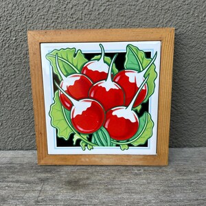 Puede incluir: Un azulejo de cerámica enmarcado con una ilustración colorida de rábanos rojos con hojas verdes sobre un fondo negro.