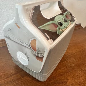 May include: A white and grey Igloo cooler with a Grogu (Baby Yoda) graphic from The Mandalorian. The cooler has a handle and a grey base. The design includes images of a spaceship.