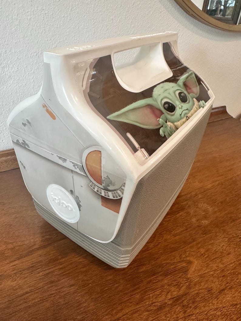 May include: A white and grey Igloo cooler with a Grogu (Baby Yoda) graphic from The Mandalorian. The cooler has a handle and a grey base. The design includes images of a spaceship.