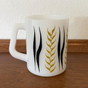 May include: A white milk glass mug featuring a repeating pattern of vertical black lines and golden-yellow wheat-like stalks. The mug has a D-shaped handle and a smooth, cylindrical body with a clean, minimalist aesthetic and a glossy finish.