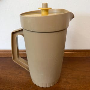 May include: A vintage tan plastic pitcher with a matching lid and a yellow handle. The pitcher has a spout for easy pouring and a handle for carrying. The lid has a yellow knob. The pitcher is sitting on a wooden surface.