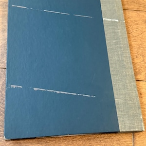 May include: A closed book with a teal blue cover and a gray spine. The cover has several white scratch marks. The spine is a light gray fabric. The book is lying on a wooden surface.