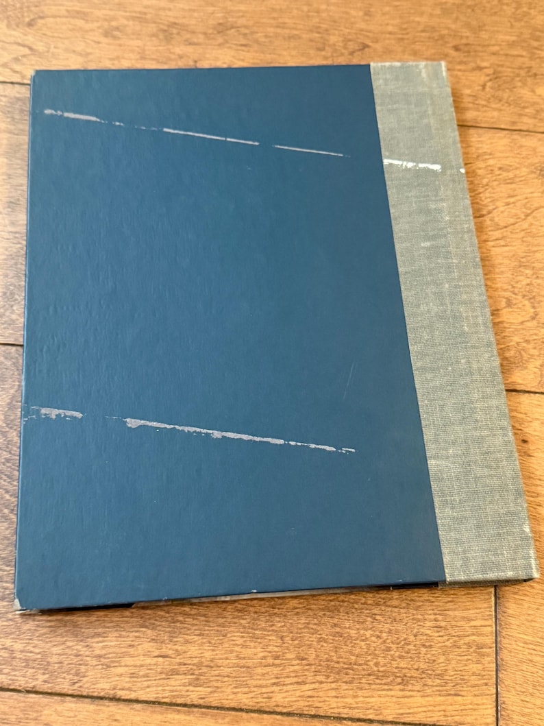 May include: A closed book with a teal blue cover and a gray spine. The cover has several white scratch marks. The spine is a light gray fabric. The book is lying on a wooden surface.