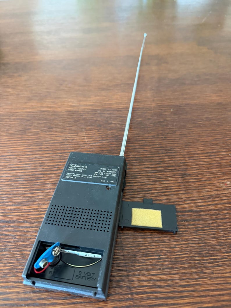 May include: A black Emerson radio with an extended silver antenna. The battery compartment is open, revealing a 9-volt battery. The radio's model number is F370, and it has a power source range.