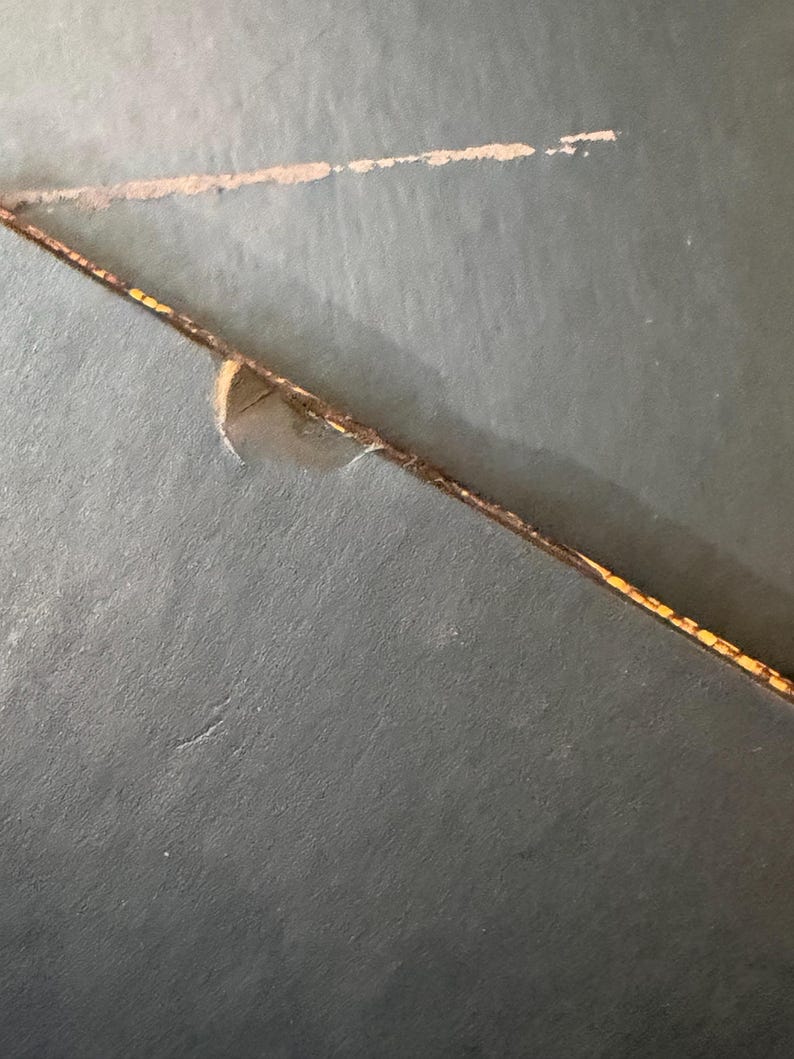 May include: Close-up of a dark teal surface with a diagonal, thin, brown line with yellow accents. A lighter, faded line is above it. A small piece of the surface is torn away.