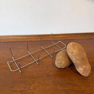 Peut inclure: Un support à pommes de terre en fil métallique argenté avec six broches verticales, à côté de deux pommes de terre brunes sur une surface en bois. Les pommes de terre sont oblongues.