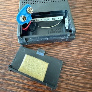 May include: Open black plastic battery compartment with a blue 9-volt battery connector and a detached cover with a yellow foam pad. The compartment has the text "9 VOLT BATTERY".