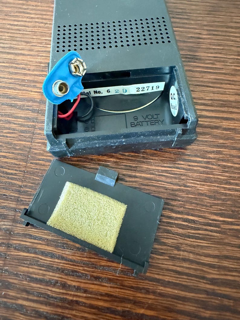 May include: Open black plastic battery compartment with a blue 9-volt battery connector and a detached cover with a yellow foam pad. The compartment has the text "9 VOLT BATTERY".
