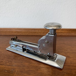 May include: A vintage silver stapler with a rounded top and a rectangular base. The stapler is made of metal and has a classic design. The stapler is sitting on a wooden surface.