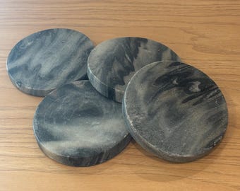 May include: Four circular coasters with a marbled grey and black design. The coasters are made from a solid material and have a smooth surface. They are arranged on a wooden surface.