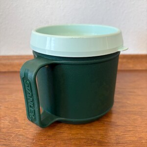 May include: A green Stanley insulated mug with a light green lid and handle. The mug is cylindrical with a handle on the side. The Stanley logo is visible on the handle. The mug is sitting on a wooden surface.