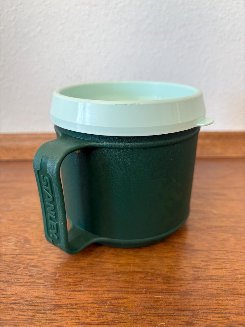 May include: A green Stanley insulated mug with a light green lid and handle. The mug is cylindrical with a handle on the side. The Stanley logo is visible on the handle. The mug is sitting on a wooden surface.