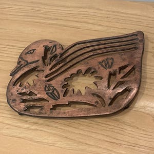 May include: A copper-coloured duck-shaped trivet with a detailed cutout design. The trivet features a duck with a detailed head, wings, and a floral and leaf pattern. The trivet is designed to protect surfaces from hot cookware.
