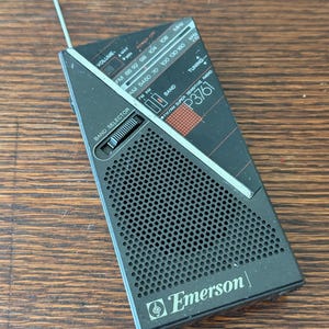 May include: Black and gray Emerson P3761 portable radio with an extendable antenna. The radio features a band selector, tuning dial, and volume control. The face of the radio has the text "FM/AM SUPER SENSITIVE RADIO".