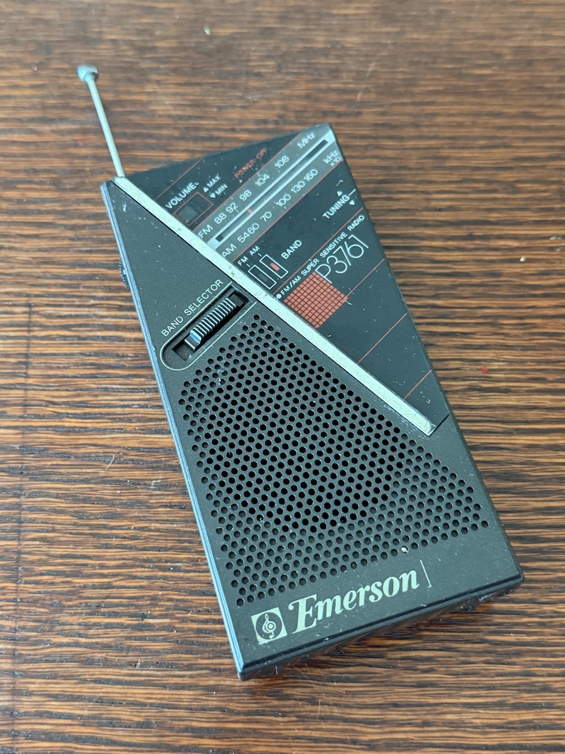 May include: Black and gray Emerson P3761 portable radio with an extendable antenna. The radio features a band selector, tuning dial, and volume control. The face of the radio has the text "FM/AM SUPER SENSITIVE RADIO".