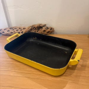 Puede incluir: Una fuente rectangular para hornear con interior negro y exterior amarillo brillante. El plato tiene dos asas amarillas a cada lado. El plato está sobre una superficie de madera.