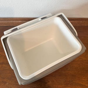 May include: A white and grey cooler with a rectangular shape. The cooler has a white interior and a grey exterior. The lid is open, revealing the inside. It is on a wooden surface.