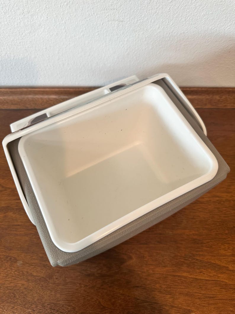 May include: A white and grey cooler with a rectangular shape. The cooler has a white interior and a grey exterior. The lid is open, revealing the inside. It is on a wooden surface.
