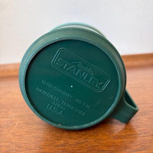 May include: Green Stanley Aladdin travel mug. The bottom of the mug is visible, showing the brand name, capacity of 12 oz, and the location of Nashville, Tennessee, USA. The mug has a handle.
