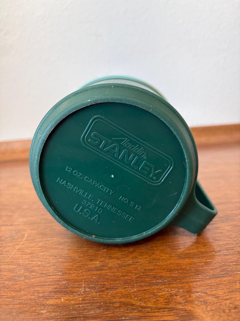 May include: Green Stanley Aladdin travel mug. The bottom of the mug is visible, showing the brand name, capacity of 12 oz, and the location of Nashville, Tennessee, USA. The mug has a handle.