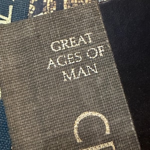 May include: Close-up of a book cover with the text "GREAT AGES OF MAN" in gold lettering. The cover has a textured, gray-brown fabric appearance. Adjacent to the book is a black, smooth surface. The image is well-lit, highlighting the details.