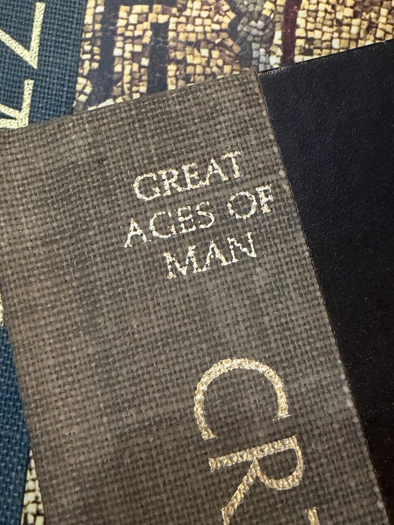 May include: Close-up of a book cover with the text "GREAT AGES OF MAN" in gold lettering. The cover has a textured, gray-brown fabric appearance. Adjacent to the book is a black, smooth surface. The image is well-lit, highlighting the details.
