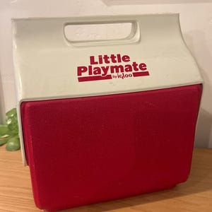May include: A white and red plastic Little Playmate by Igloo cooler with a top handle. The front features the text Little Playmate by igloo in red lettering. The cooler has a rectangular shape with a hinged lid design and a textured red front panel.