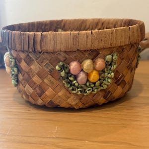 以下が含まれることがあります： 装飾的な花の模様の丸い織りバスケット。バスケットは、天然色の織り素材で作られており、上部に装飾的なトリムが付いています。花の模様は、ピンク、黄色、オレンジ色のアクセントが特徴です。