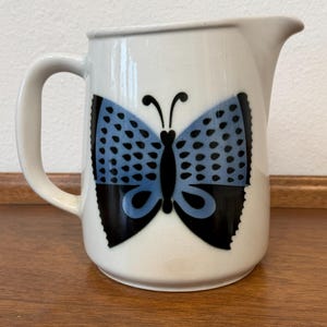 May include: White ceramic pitcher with a black handle and spout. The pitcher features a large butterfly design in black and blue on the front. The butterfly has a detailed wing pattern. The pitcher is likely used for serving beverages.