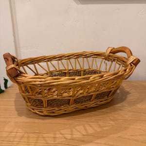 May include: An oval-shaped woven basket featuring light brown willow reeds and a darker brown woven base. The basket includes two sturdy wooden handles attached to the sides, with an open-weave design along the upper rim and a solid base.