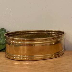 May include: A gold-colored, oval-shaped planter. The planter has a series of horizontal ridges around its exterior. The interior is a darker gold color. The planter is sitting on a wooden surface.