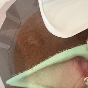 May include: Close-up of a section of a Star Wars-themed image. The image features a brown, textured surface, a light green ear, and a portion of a white object. The ear has a pink interior. The image is partially obscured by a white, curved object.