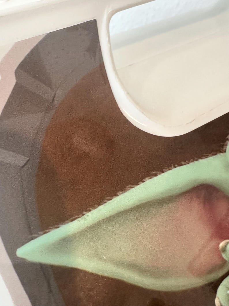 May include: Close-up of a section of a Star Wars-themed image. The image features a brown, textured surface, a light green ear, and a portion of a white object. The ear has a pink interior. The image is partially obscured by a white, curved object.