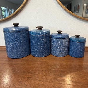 Peut inclure: Un ensemble de quatre boîtes cylindriques avec une finition émaillée mouchetée bleue et blanche. Chaque boîte possède un couvercle assorti surmonté d'un petit bouton circulaire marron foncé. Les boîtes sont alignées par taille décroissante.