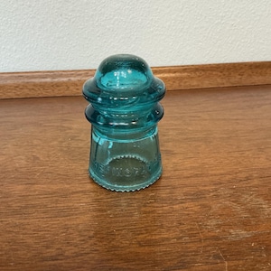 Può includere: Un isolatore in vetro turchese vintage con una parte superiore arrotondata e una base svasata. L'isolatore ha un design a strati ed è su una superficie di legno. Il testo "HEMINGRAY" è visibile sul lato del vetro.