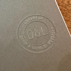 May include: Close-up of a gray paper with a circular embossed design. The design reads "LIBRARY OF DAVID R. LOTEL&Eacute;I WOOD" around the perimeter, with "D & L" in the center. The paper has a textured surface and a white border.