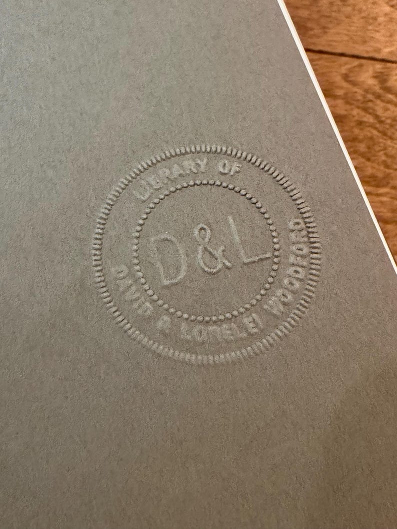 May include: Close-up of a gray paper with a circular embossed design. The design reads "LIBRARY OF DAVID R. LOTEL&Eacute;I WOOD" around the perimeter, with "D & L" in the center. The paper has a textured surface and a white border.