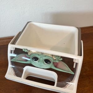 May include: A white and grey plastic storage container with a Grogu (Baby Yoda) graphic from The Mandalorian. The container is rectangular with a handle and a clear plastic front.