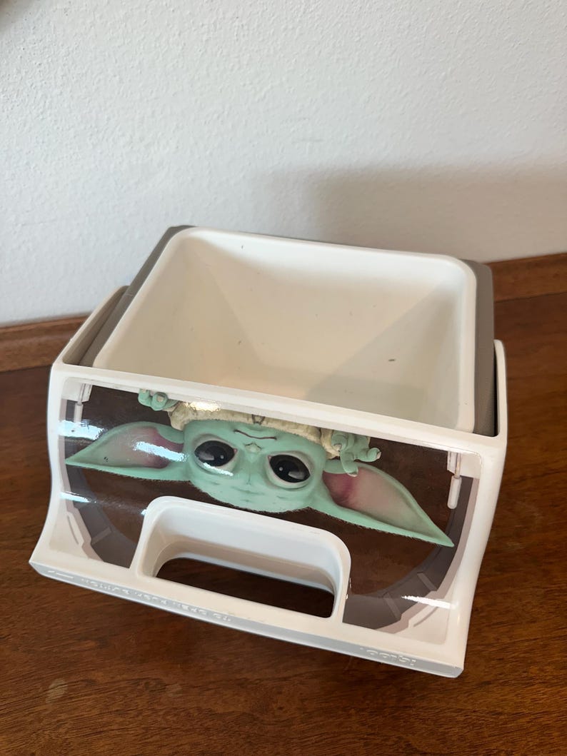 May include: A white and grey plastic storage container with a Grogu (Baby Yoda) graphic from The Mandalorian. The container is rectangular with a handle and a clear plastic front.