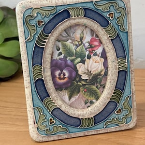 May include: An oval picture frame with a floral print. The frame has a blue and cream-colored border with decorative accents. The picture inside features a bouquet of flowers, including roses and pansies.