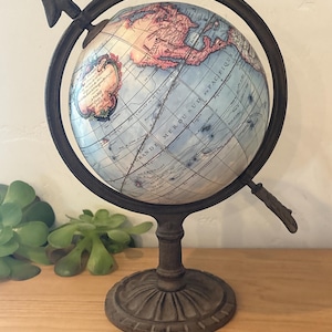May include: A decorative globe featuring a vintage-style map with light blue oceans and tan landmasses. The globe is mounted in a dark brown, weathered metal frame with a circular base and an arrow-shaped pointer at the top, displaying French text.