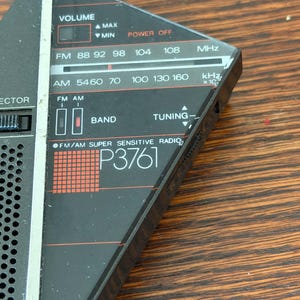 May include: A vintage black and silver FM/AM radio with a tuning dial and frequency display. The radio features a volume control, band selection, and the text "P3761". The radio's frequency range is displayed in MHz and kHz.