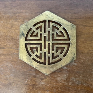 Puede incluir: Posavasos hexagonal de latón con diseño geométrico. El posavasos presenta un patrón laberíntico recortado en el centro. El latón tiene un aspecto ligeramente envejecido, con algunas manchas más oscuras. El posavasos está sobre una superficie de madera.