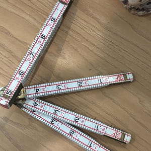 May include: A white and red folding ruler with black numbers and markings. The ruler is made of wood and has brass hinges. The ruler is partially open, displaying measurements in inches and feet. The ruler is lying on a wooden surface.