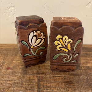 Puede incluir: Un par de saleros y pimenteros de cerámica marrón con diseños florales. Un salero tiene una flor blanca con hojas verdes y el otro tiene una flor amarilla con hojas verdes.