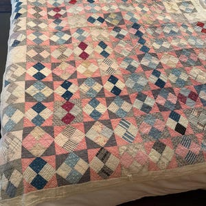May include: A patchwork quilt featuring a repeating block pattern composed of various fabric squares in shades of pink, blue, grey, and white. The textile displays a grid of alternating solid and patterned squares with visible stitching throughout the surface.